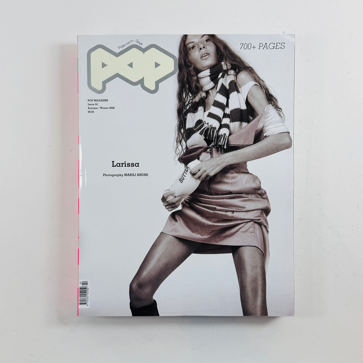A single magazine or book cover titled "POP" lies flat against a plain light background. The cover features a grayscale fashion photo of a woman wearing a striped scarf and a metallic pink mini dress, with text including "700+ PAGES" and the name "Larissa.