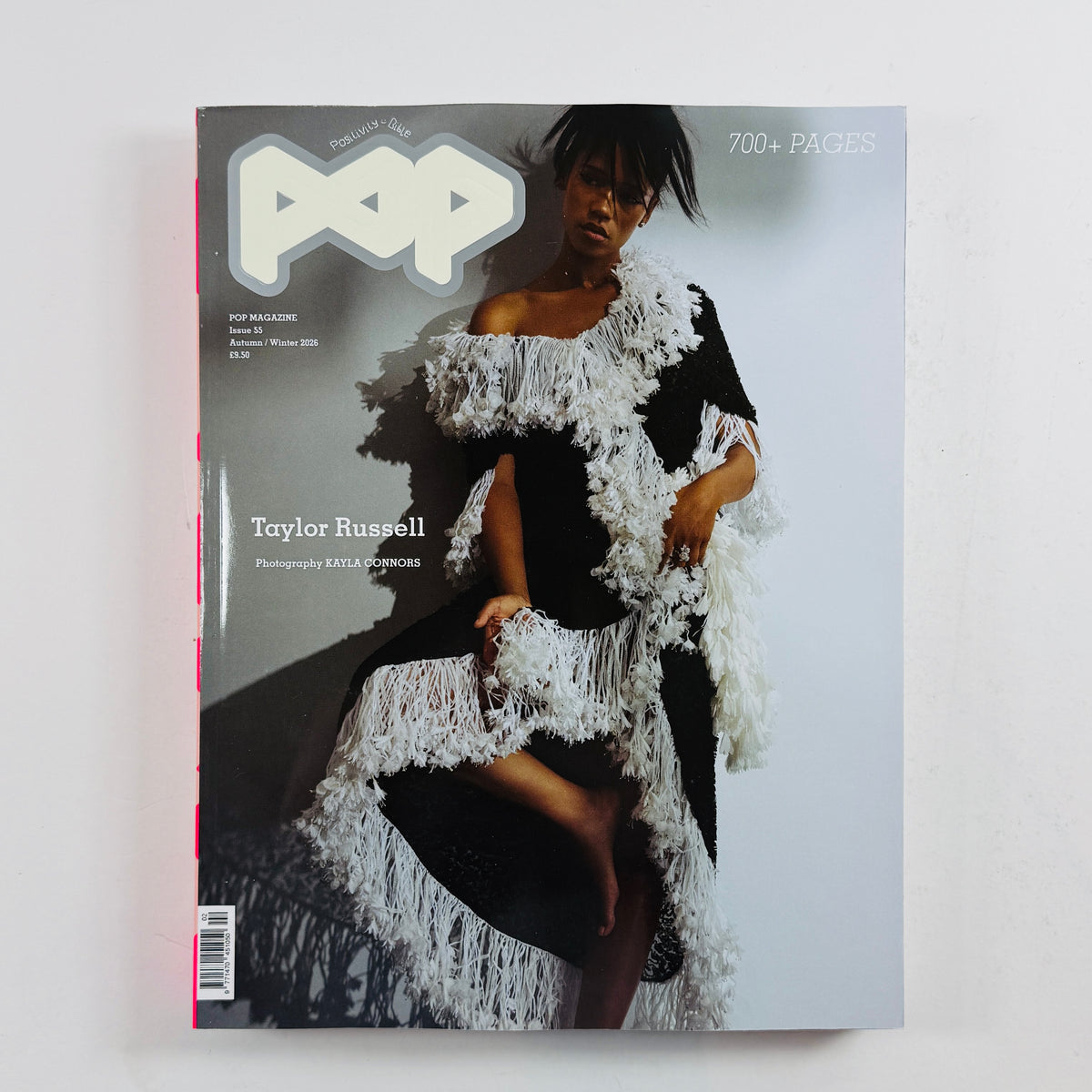 A magazine titled 'POP' lying flat on a light gray surface. The cover features a fashion photograph of a woman wearing a black dress with dramatic white feathered or fringed trim, and the cover text includes 'Taylor Russell' and '700+ PAGES'.